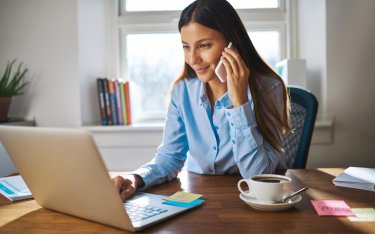 Eine Frau in einem blauen Hemd sitzt an einem Schreibtisch, telefoniert und arbeitet an einem Laptop, in der Nähe stehen eine Tasse Kaffee und Haftnotizen.
