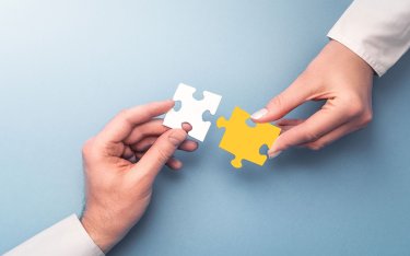 Zwei Hände halten weiße und gelbe Puzzlestücke, die sie vor einem hellblauen Hintergrund so positionieren, dass sie zusammenpassen.