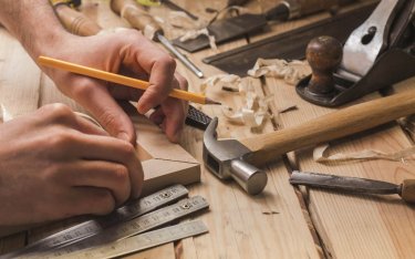 Eine Person, die mit einem Bleistift ein Stück Holz auf einer Werkbank anzeichnet, umgeben von Tischlerwerkzeugen wie Hammer, Lineal, Meißel und Holzspänen.