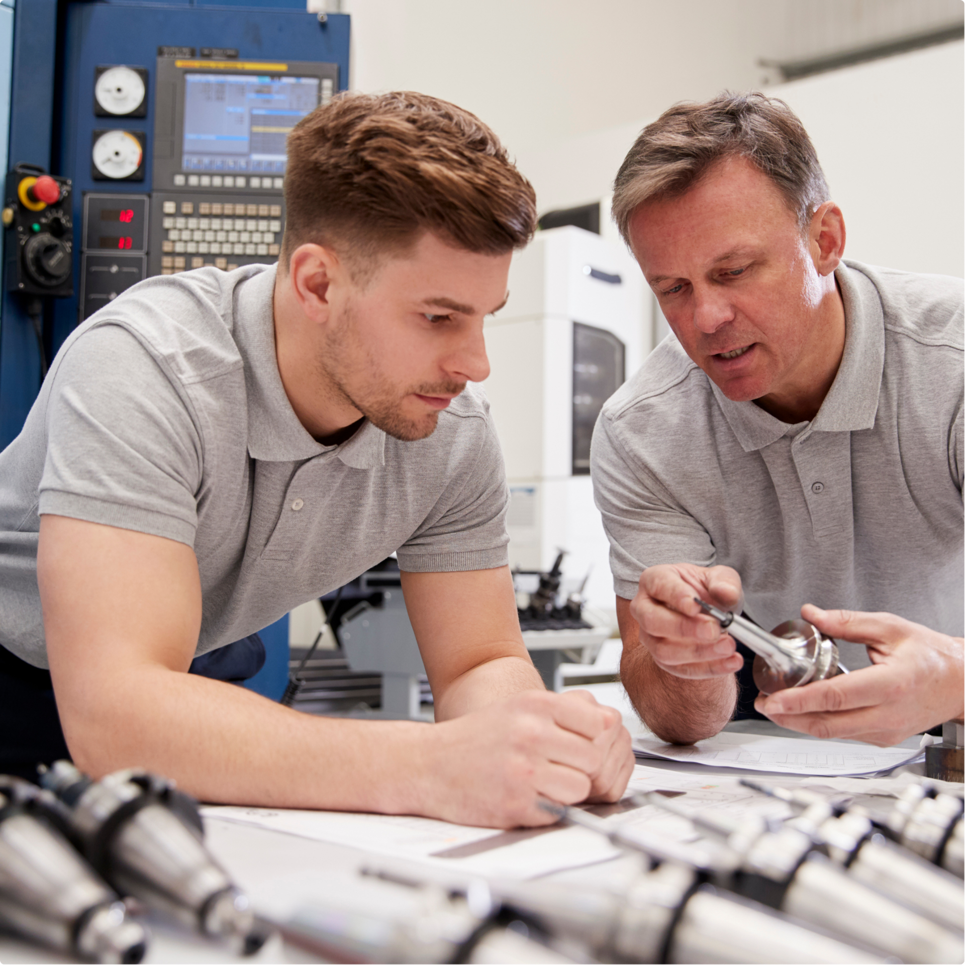 Zwei Männer in grauen Polohemden untersuchen gemeinsam ein Maschinenteil aus Metall in einer Industriehalle, im Hintergrund sind technische Diagramme und Geräte zu sehen.